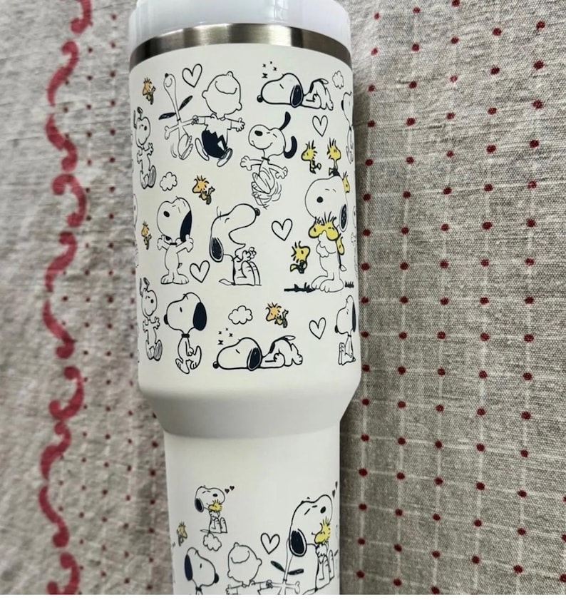 Puede incluir: Un vaso t&eacute;rmico blanco con un patr&oacute;n repetido de los personajes de Snoopy y Woodstock en negro y amarillo. El vaso tiene un borde de acero inoxidable y una tapa blanca. El dise&ntilde;o incluye corazones y otros gr&aacute;ficos peque&ntilde;os.