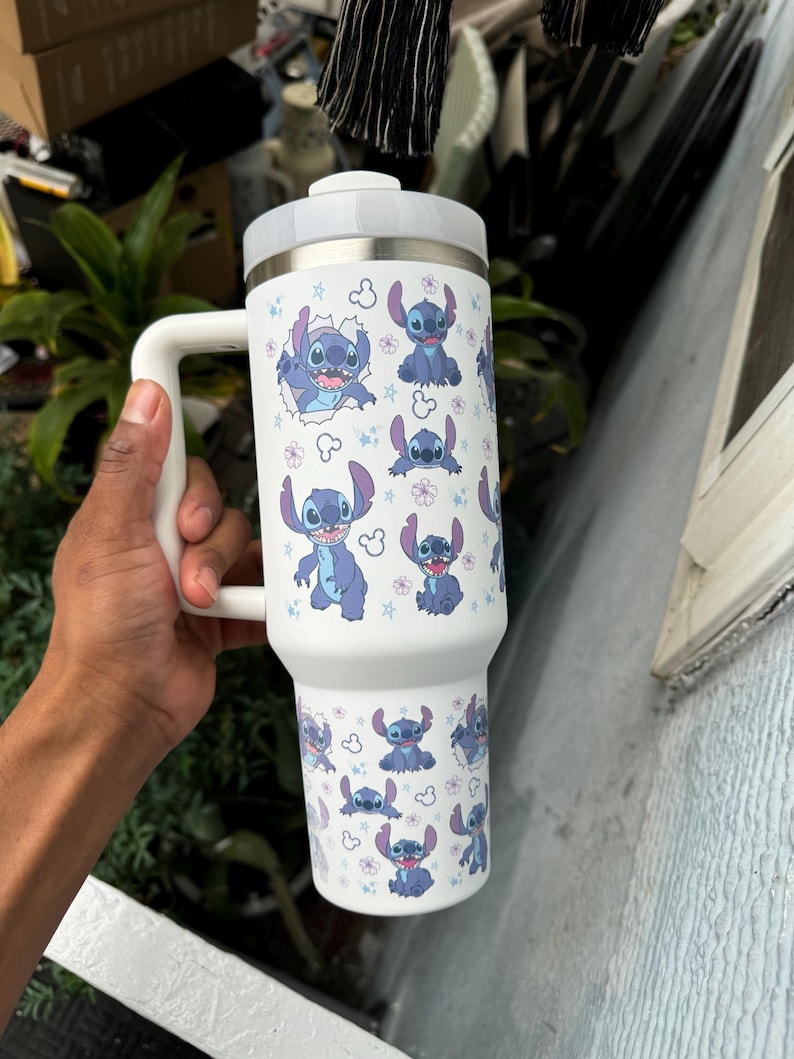May include: A white insulated tumbler with a handle, featuring a repeating pattern of the character Stitch from Lilo & Stitch. The design includes Stitch in various poses, along with flowers and stars. The tumbler has a white lid and handle.