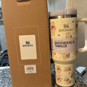 May include: A Stanley Quencher H2.0 tumbler with a Winnie the Pooh design. The 40 oz tumbler has a light yellow background with illustrations of Winnie the Pooh and other graphics. The tumbler is next to a brown cardboard box.