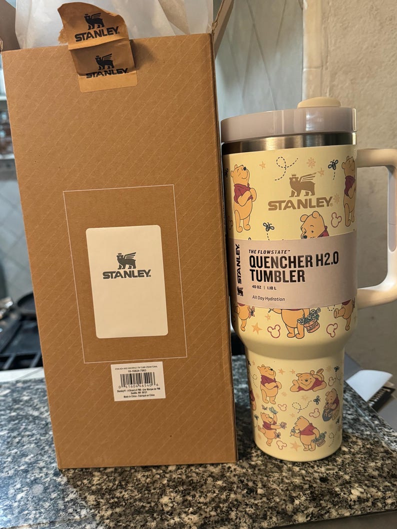 May include: A Stanley Quencher H2.0 tumbler with a Winnie the Pooh design. The 40 oz tumbler has a light yellow background with illustrations of Winnie the Pooh and other graphics. The tumbler is next to a brown cardboard box.