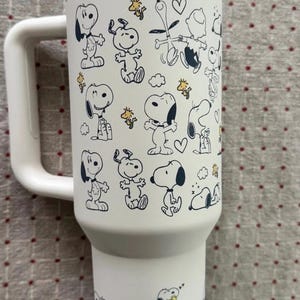 Puede incluir: Vaso t&eacute;rmico blanco con un asa grande, con un patr&oacute;n repetido de los personajes de dibujos animados Snoopy y Woodstock en negro y amarillo. El vaso tiene un borde plateado y un asa blanca.