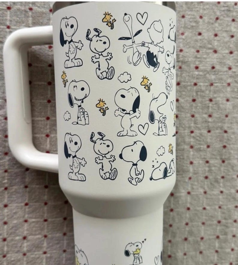 Puede incluir: Vaso t&eacute;rmico blanco con un asa grande, con un patr&oacute;n repetido de los personajes de dibujos animados Snoopy y Woodstock en negro y amarillo. El vaso tiene un borde plateado y un asa blanca.