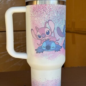 May include: White travel mug with a gray lid and handle. The mug features a colorful design of cartoon characters Stitch and Angel, with a pink and blue speckled background. The mug is designed to keep beverages hot or cold.