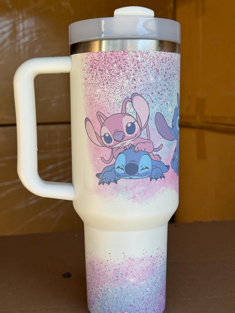 May include: White travel mug with a gray lid and handle. The mug features a colorful design of cartoon characters Stitch and Angel, with a pink and blue speckled background. The mug is designed to keep beverages hot or cold.