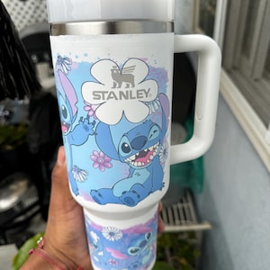 May include: White Stanley travel mug featuring a blue cartoon character design with flowers. The mug has a white handle and lid, and the Stanley logo is visible. The mug is designed to hold a large quantity of liquid.