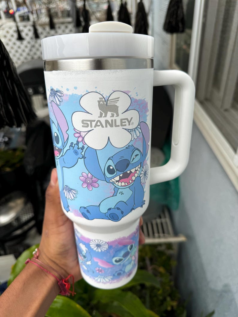 May include: White Stanley travel mug featuring a blue cartoon character design with flowers. The mug has a white handle and lid, and the Stanley logo is visible. The mug is designed to hold a large quantity of liquid.