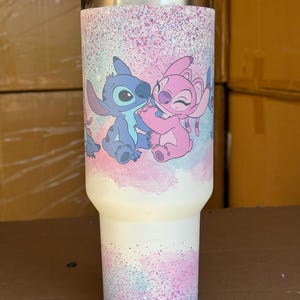 May include: A white insulated tumbler featuring a colorful design of the characters Stitch and Angel from the animated movie Lilo & Stitch. The design includes a pink and blue speckled background. The tumbler has a silver lid.