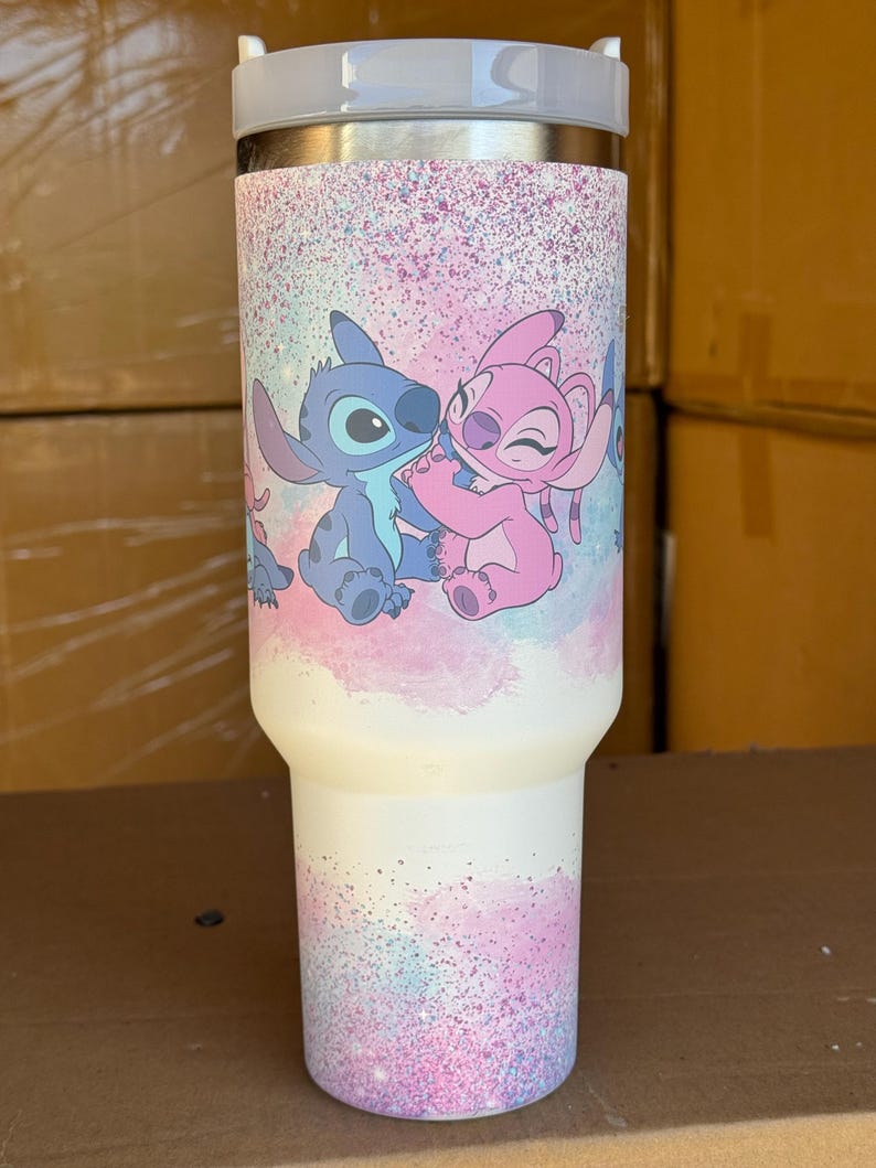 May include: A white insulated tumbler featuring a colorful design of the characters Stitch and Angel from the animated movie Lilo & Stitch. The design includes a pink and blue speckled background. The tumbler has a silver lid.