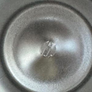 Puede incluir: Primer plano de un objeto met&aacute;lico circular con un componente central de pl&aacute;stico transparente elevado. La superficie tiene un aspecto de metal cepillado con sutiles reflejos de luz. El dise&ntilde;o del objeto sugiere un componente funcional, posiblemente mec&aacute;nico.