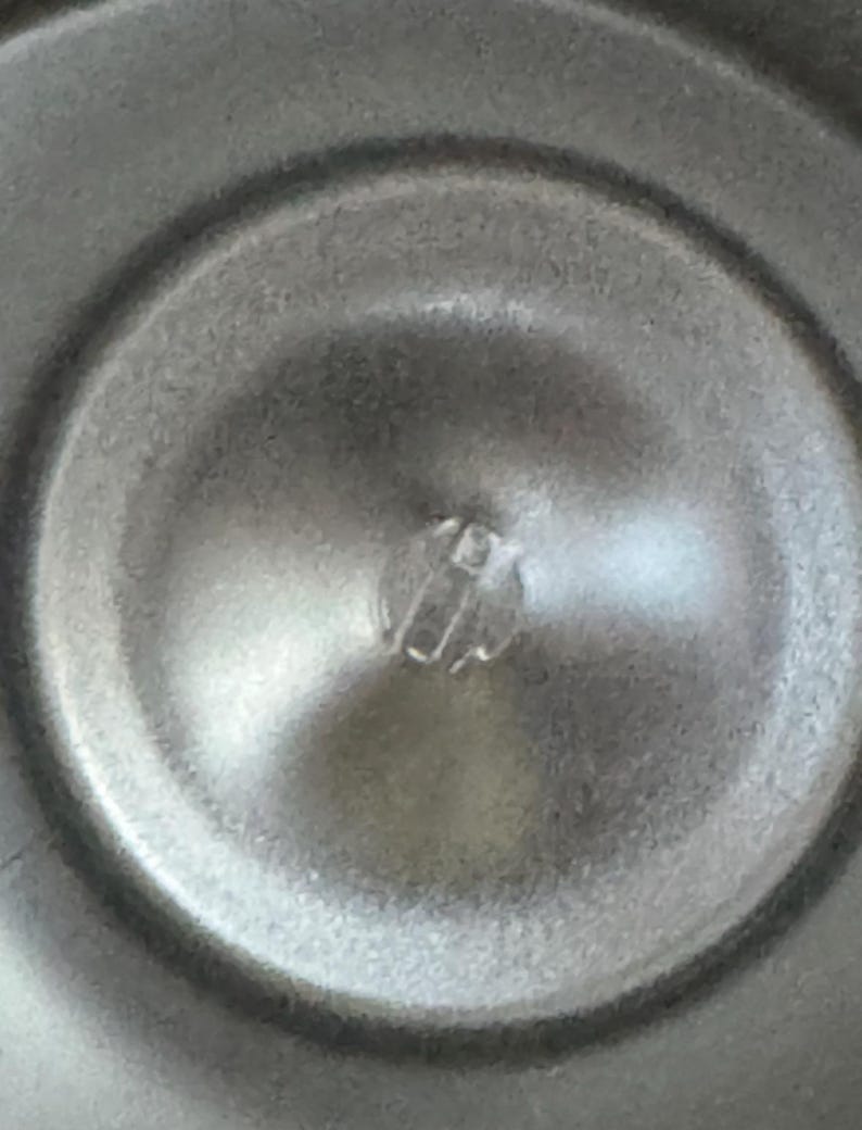 Puede incluir: Primer plano de un objeto met&aacute;lico circular con un componente central de pl&aacute;stico transparente elevado. La superficie tiene un aspecto de metal cepillado con sutiles reflejos de luz. El dise&ntilde;o del objeto sugiere un componente funcional, posiblemente mec&aacute;nico.