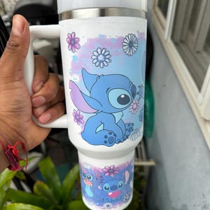 May include: A white travel mug featuring a blue cartoon character with large eyes and a floral design. The mug has a white handle and lid, and a stainless steel rim. The design includes pink and blue flowers.
