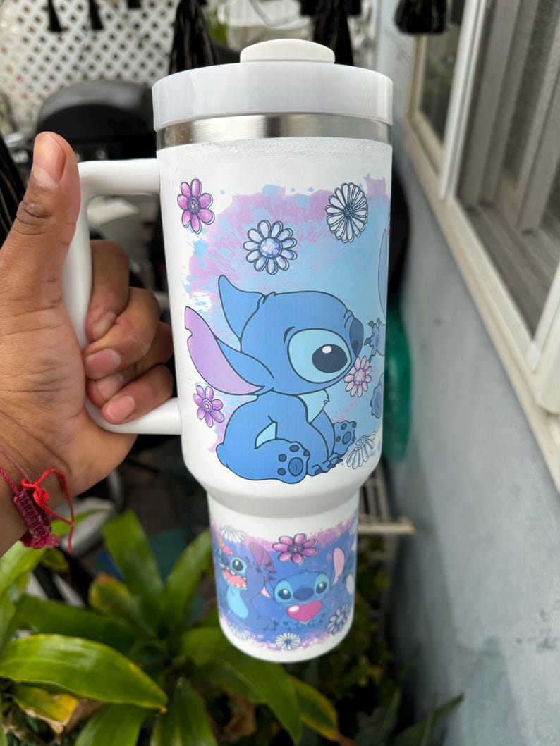 May include: A white travel mug featuring a blue cartoon character with large eyes and a floral design. The mug has a white handle and lid, and a stainless steel rim. The design includes pink and blue flowers.