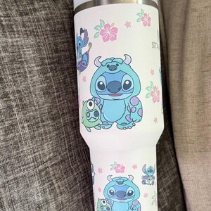Puede incluir: Un vaso t&eacute;rmico blanco con los personajes de dibujos animados Stitch y Sulley, junto con flores de hibisco rosas y estrellas. El vaso tiene una tapa azul claro y un borde plateado. El dise&ntilde;o se repite alrededor del cuerpo del vaso.