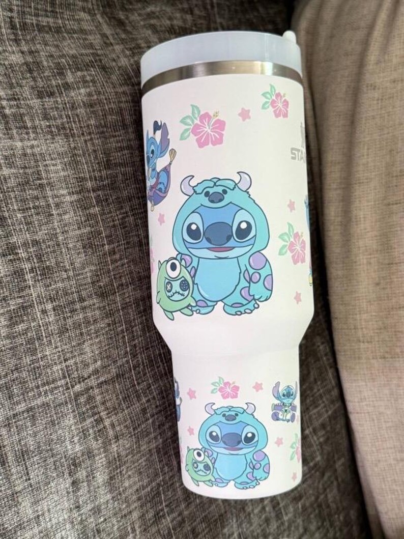 Puede incluir: Un vaso t&eacute;rmico blanco con los personajes de dibujos animados Stitch y Sulley, junto con flores de hibisco rosas y estrellas. El vaso tiene una tapa azul claro y un borde plateado. El dise&ntilde;o se repite alrededor del cuerpo del vaso.