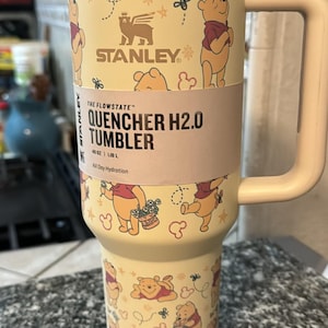 May include: A Stanley Quencher H2.0 tumbler with a Winnie the Pooh theme. The light yellow tumbler has a handle and a lid. The design features Winnie the Pooh and other graphics. The tumbler holds 40 oz (1.18 L).