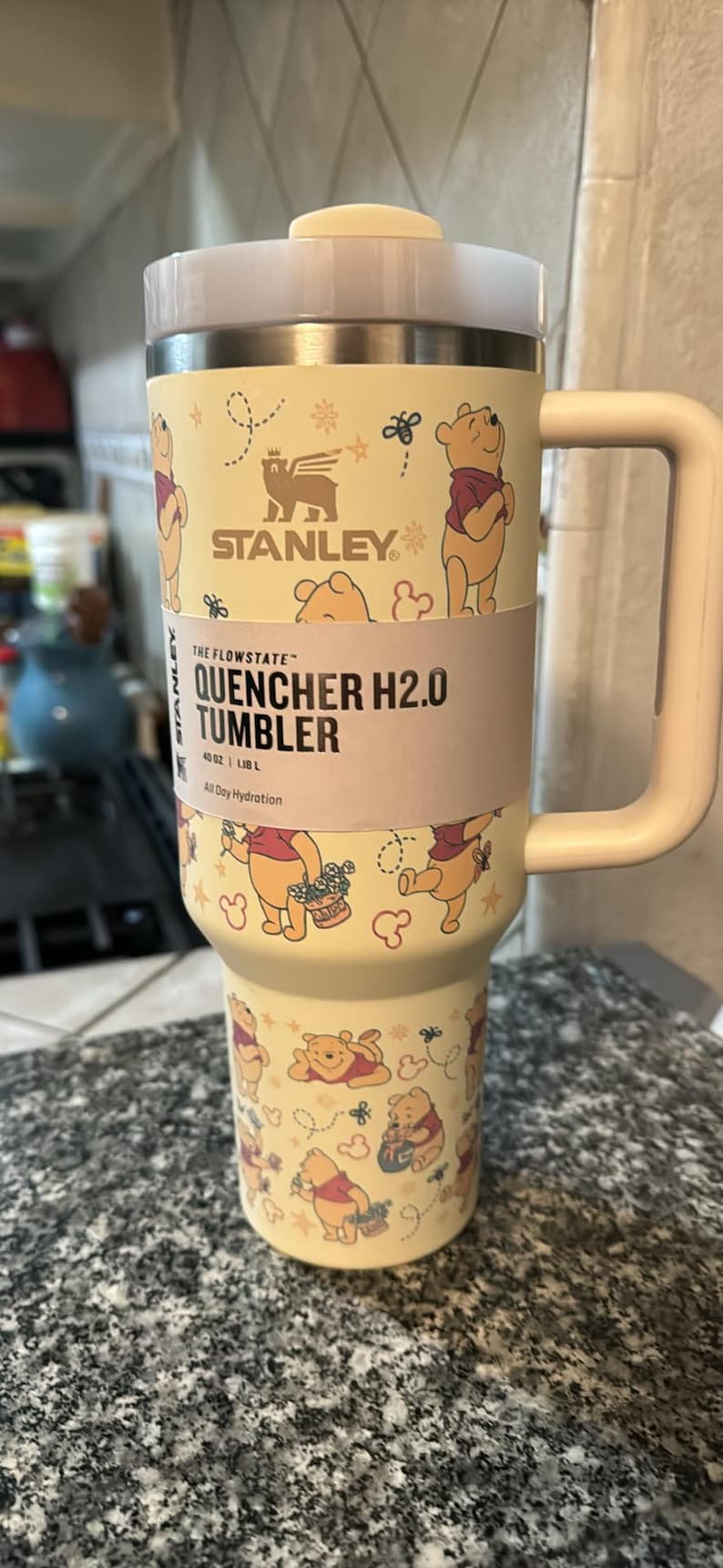 May include: A Stanley Quencher H2.0 tumbler with a Winnie the Pooh theme. The light yellow tumbler has a handle and a lid. The design features Winnie the Pooh and other graphics. The tumbler holds 40 oz (1.18 L).