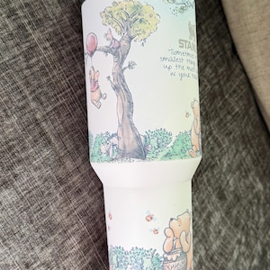 May include: A white insulated tumbler featuring a whimsical illustration of Winnie the Pooh and friends. The design includes a tree, a red balloon, and the quote, "Sometimes the smallest things take up the most room in your heart."