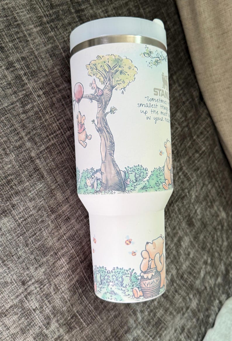 May include: A white insulated tumbler featuring a whimsical illustration of Winnie the Pooh and friends. The design includes a tree, a red balloon, and the quote, "Sometimes the smallest things take up the most room in your heart."