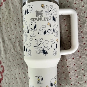 Puede incluir: Taza de viaje Stanley blanca con ilustraciones de dibujos animados de Snoopy y Woodstock. La taza tiene un asa blanca y un borde plateado. El logotipo de Stanley est&aacute; impreso en la parte delantera. Para bebidas fr&iacute;as o calientes.