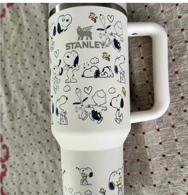Puede incluir: Taza de viaje Stanley blanca con ilustraciones de dibujos animados de Snoopy y Woodstock. La taza tiene un asa blanca y un borde plateado. El logotipo de Stanley est&aacute; impreso en la parte delantera. Para bebidas fr&iacute;as o calientes.