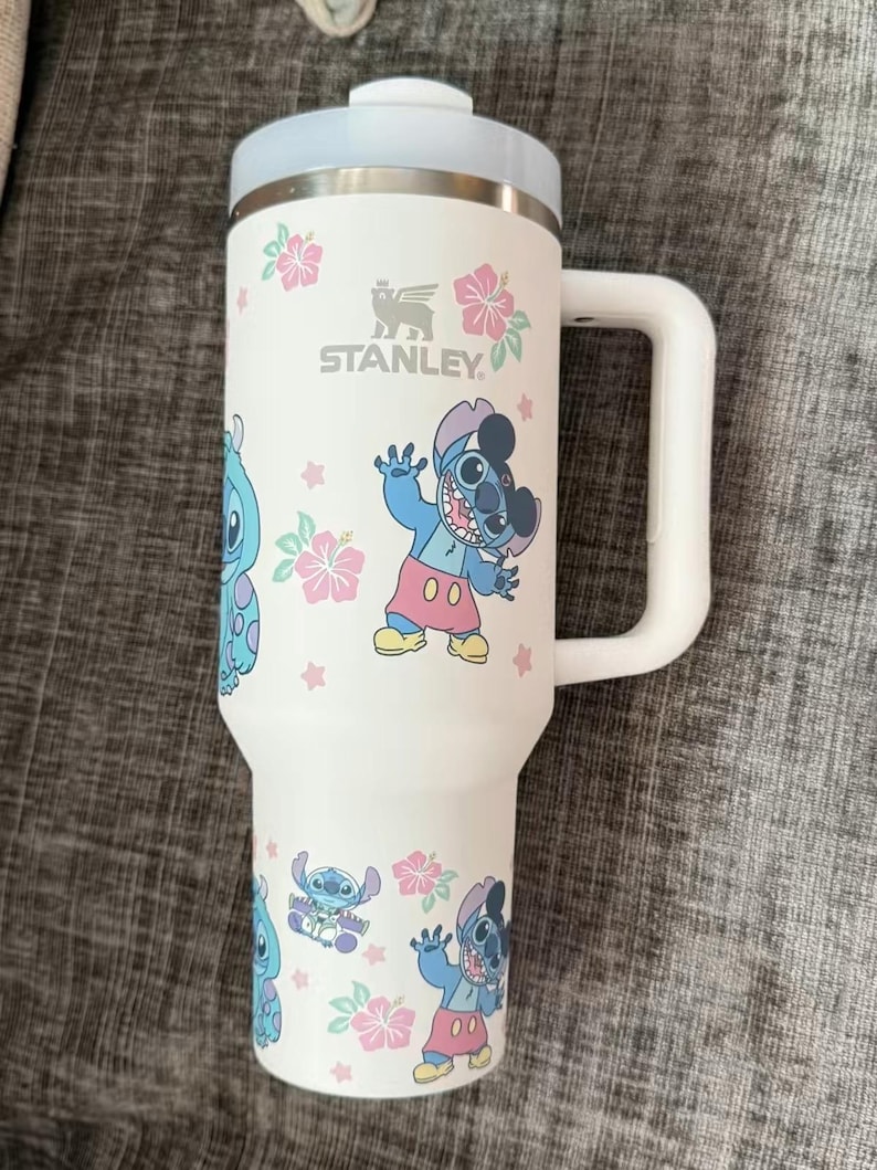 Puede incluir: Taza de viaje Stanley blanca con un dise&ntilde;o de dibujos animados de Stitch, flores rosas y estrellas. La taza tiene un asa blanca, una tapa azul claro y el logotipo de Stanley.