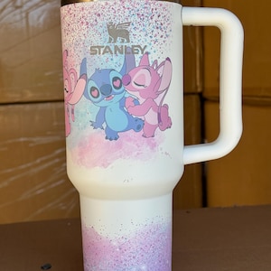May include: White Stanley tumbler with a handle, featuring a colorful design of cartoon characters. The design includes the Stanley logo and a pastel glitter effect. The tumbler has a white lid and handle.