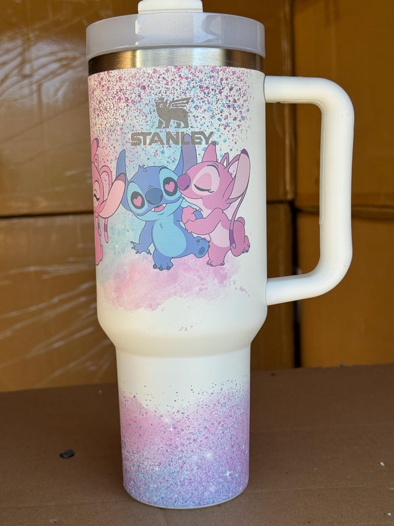 May include: White Stanley tumbler with a handle, featuring a colorful design of cartoon characters. The design includes the Stanley logo and a pastel glitter effect. The tumbler has a white lid and handle.