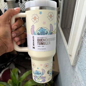 Puede incluir: Vaso Stanley Quencher H2.0 color crema con asa y tapa. El vaso presenta un dise&ntilde;o de personaje de dibujos animados con flores y estrellas. El texto "THE FLOWSTATE QUENCHER H2.0 TUMBLER" est&aacute; impreso en el lateral. Contiene 1,18 L.