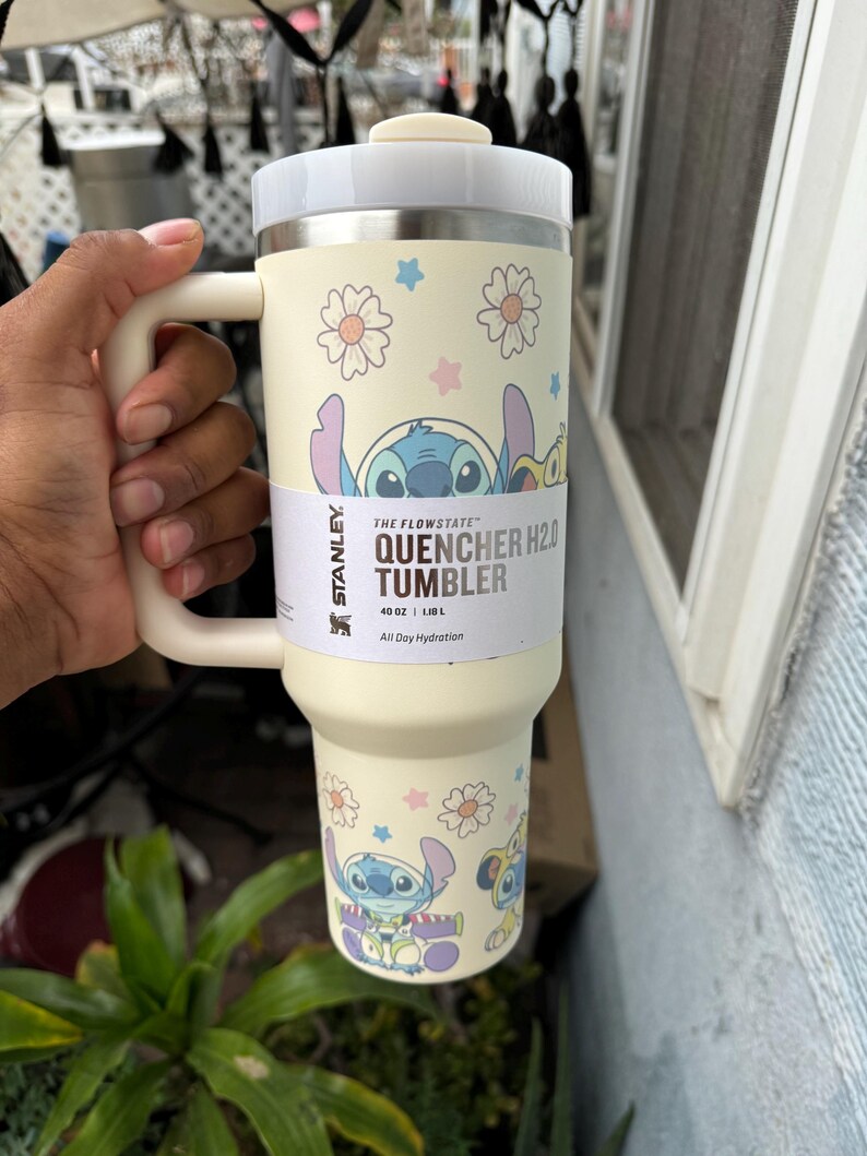 Puede incluir: Vaso Stanley Quencher H2.0 color crema con asa y tapa. El vaso presenta un dise&ntilde;o de personaje de dibujos animados con flores y estrellas. El texto "THE FLOWSTATE QUENCHER H2.0 TUMBLER" est&aacute; impreso en el lateral. Contiene 1,18 L.