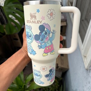 Puede incluir: Vaso Stanley crema con asa blanca, con personajes de dibujos animados y dise&ntilde;os florales. El vaso tiene el logotipo de Stanley y presenta im&aacute;genes de Stitch y otros personajes. El vaso est&aacute; dise&ntilde;ado para contener una gran cantidad de l&iacute;quido.