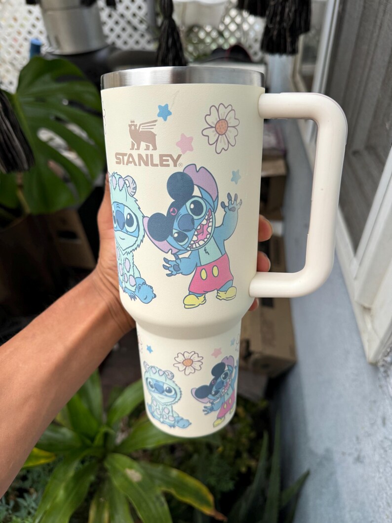 Puede incluir: Vaso Stanley crema con asa blanca, con personajes de dibujos animados y dise&ntilde;os florales. El vaso tiene el logotipo de Stanley y presenta im&aacute;genes de Stitch y otros personajes. El vaso est&aacute; dise&ntilde;ado para contener una gran cantidad de l&iacute;quido.