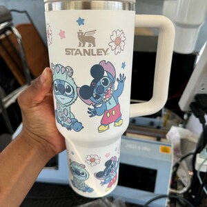 Puede incluir: Vaso Stanley blanco con asa, con el personaje de dibujos animados Stitch y dise&ntilde;os florales. El vaso tiene tapa y asa blancas, con el logotipo de Stanley. El dise&ntilde;o incluye detalles en azul, rosa y amarillo.