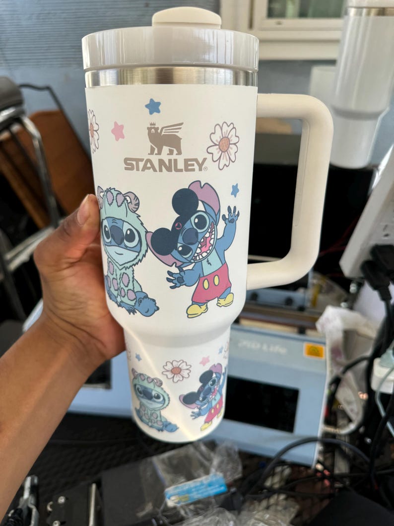 Puede incluir: Vaso Stanley blanco con asa, con el personaje de dibujos animados Stitch y dise&ntilde;os florales. El vaso tiene tapa y asa blancas, con el logotipo de Stanley. El dise&ntilde;o incluye detalles en azul, rosa y amarillo.