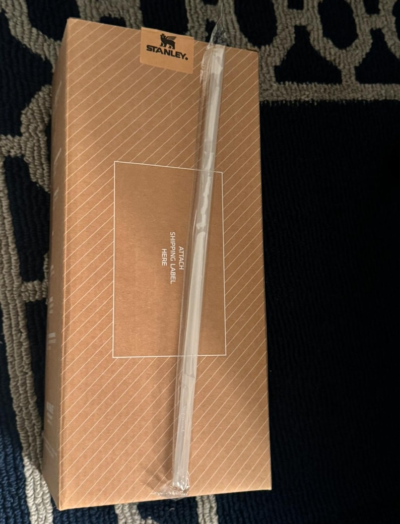 Puede incluir: Una caja de cart&oacute;n marr&oacute;n con rayas diagonales. La caja tiene una correa de pl&aacute;stico transparente y una etiqueta que dice "STANLEY". Se adjunta una etiqueta transparente con el texto "ATTACH SHIPPING LABEL HERE."