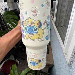 Puede incluir: Un vaso de color crema con personajes de dibujos animados y dise&ntilde;os florales. El vaso tiene un borde de acero inoxidable y est&aacute; decorado con im&aacute;genes de un le&oacute;n amarillo y un extraterrestre azul. El dise&ntilde;o incluye flores rosas y azules.