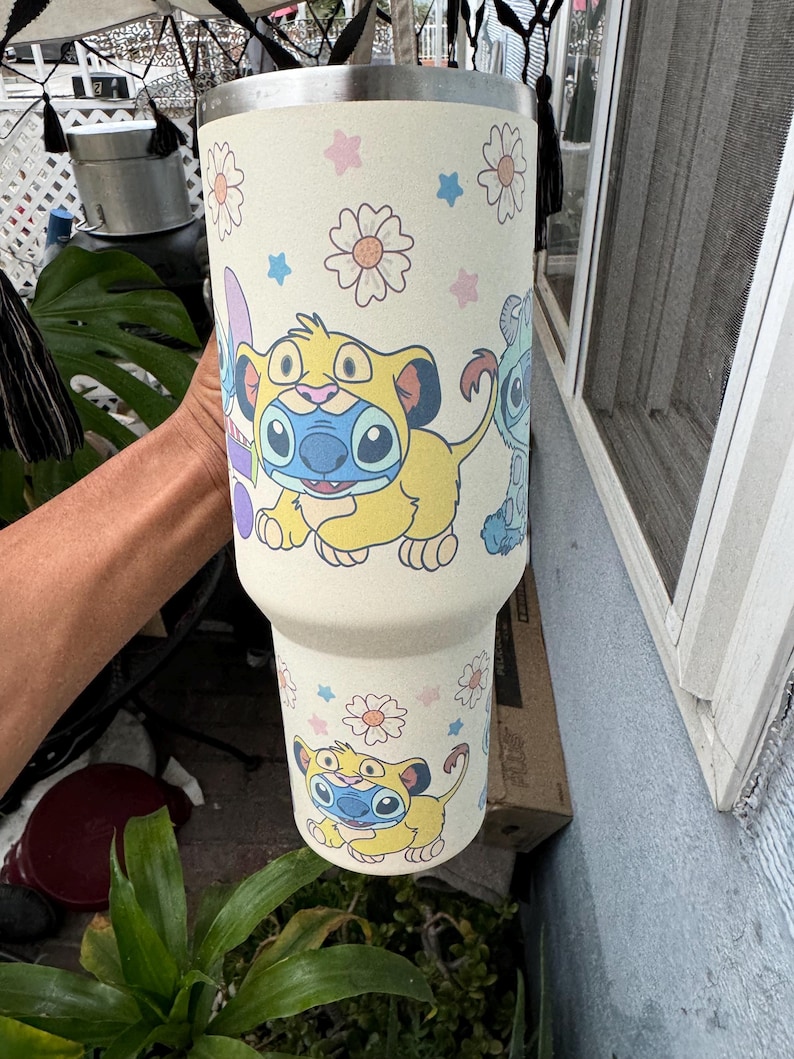 Puede incluir: Un vaso de color crema con personajes de dibujos animados y dise&ntilde;os florales. El vaso tiene un borde de acero inoxidable y est&aacute; decorado con im&aacute;genes de un le&oacute;n amarillo y un extraterrestre azul. El dise&ntilde;o incluye flores rosas y azules.