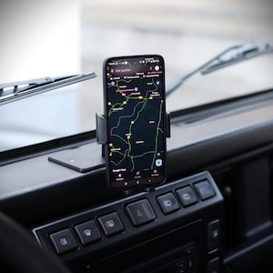 Könnte beinhalten: Ein schwarzes Smartphone mit einer Kartenanwendung ist in einem Auto montiert. Das Telefon wird von einer schwarzen Telefonhalterung am Armaturenbrett gehalten. Das Armaturenbrett hat mehrere Knöpfe und einen Scheibenwischer.
