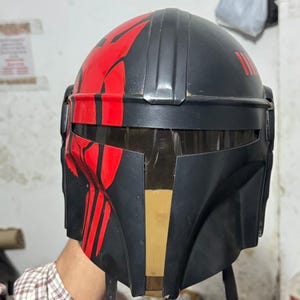 May include: A black helmet with red accents, featuring a distinctive design on the front and top. The helmet has a visor and straps, and appears to be a costume piece or prop.