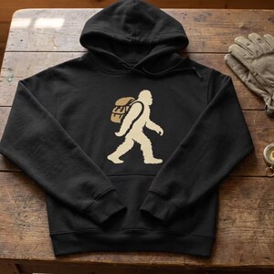 May include: Black hooded sweatshirt with a Bigfoot graphic in beige and tan. The design features a silhouette of Bigfoot carrying a backpack. The sweatshirt is on a wooden surface with gloves and a compass.