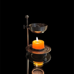 May include: A dark metal oil burner featuring an adjustable vertical stand, a circular base with an embossed pattern, and a top bowl. A bright orange candle sits on the base, providing light to the bowl above against a black background.