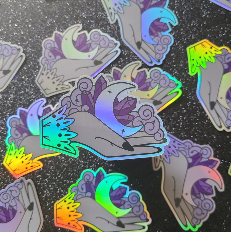 Puede incluir: Un conjunto de pegatinas hologr&aacute;ficas iridiscentes con una mano sosteniendo una luna creciente y cristales. Las pegatinas tienen un efecto arco&iacute;ris y son perfectas para agregar un toque m&iacute;stico a tus diarios, port&aacute;tiles o botellas de agua.