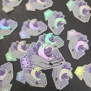 Puede incluir: Un conjunto de pegatinas hologr&aacute;ficas iridiscentes con una mano sosteniendo una luna creciente y cristales. Las pegatinas son perfectas para agregar un toque de magia a tu agenda, computadora port&aacute;til o botella de agua.