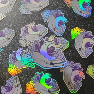 Puede incluir: Un conjunto de pegatinas hologr&aacute;ficas iridiscentes con una mano sosteniendo una luna creciente y cristales. Las pegatinas son perfectas para agregar un toque de magia a tus diarios, port&aacute;tiles, botellas de agua y m&aacute;s.