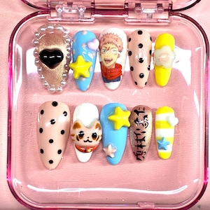 May include: A set of ten press-on nails in a pink case, featuring varied designs including a gold nail with a black heart and pearls, blue nails with yellow stars, a nail with an anime character, polka dots, a cat face, and Japanese text.