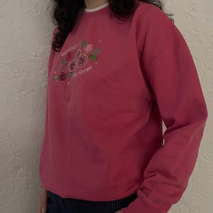 Könnte beinhalten: Ein magentafarbenes Sweatshirt mit weißem Kragen und gesticktem Blumendesign. Der Text "Grandma's" und "us forever" ist über den Blumen gestickt. Das Sweatshirt hat lange Ärmel und gerippte Bündchen.