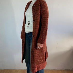 Könnte beinhalten: Ein langer, rostfarbener Strickmantel mit Knopfleiste. Der Mantel hat ein strukturiertes Aussehen und ein klassisches, maßgeschneidertes Design. Die Ärmel sind lang und der Mantel reicht bis zur Mitte der Wade. Der Mantel wird über einem weißen Rollkragenpullover und einer blauen Jeans getragen.