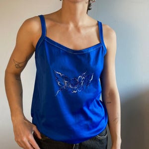 Könnte beinhalten: Ein leuchtend blaues Tanktop mit dünnen Trägern und weißer Naht. Das Oberteil hat eine detaillierte Blumenstickerei in Weiß- und Silbertönen auf der Vorderseite. Das Tanktop wird mit einer dunklen Jeans kombiniert.