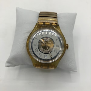 Könnte beinhalten: Eine Vintage-Swatch-Uhr mit transparentem gelbem Kunststoffgehäuse und einem goldfarbenen Metallarmband. Das Zifferblatt ist weiß mit schwarzen römischen Ziffern und goldfarbenen Zeigern. Das Datum wird auf 3 Uhr angezeigt.