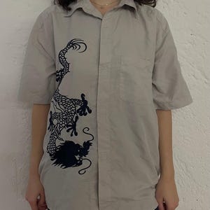 Könnte beinhalten: Hellgraues Hemd mit kurzen Ärmeln und einem schwarzen Drachendesign auf der linken Seite. Das Hemd hat eine Knopfleiste, eine Tasche und einen klassischen Kragen. Hergestellt aus leichtem Stoff, ideal für den täglichen Gebrauch.