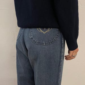 Könnte beinhalten: Eine Person trägt einen marineblauen Pullover und eine hellblaue Jeans. Die Jeans hat ein dekoratives Design auf der Gesäßtasche. Die Person trägt mehrere Ringe.
